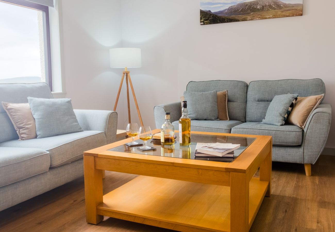 Comfortable living room with grey sofas, wooden coffee table set with whisky, tripod lamp and Highland landscape artwork at Lynemore Croft