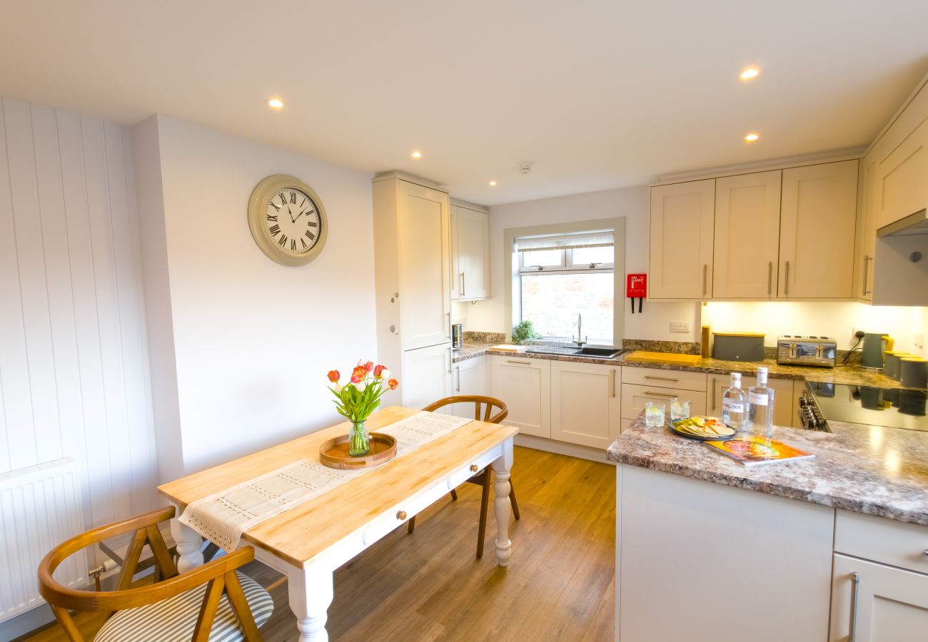 Dining area with white farmhouse table, fresh tulips and wall clock adjacent to kitchen at Lynemore Croft