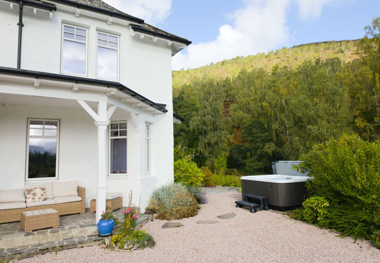 White house with covered veranda, rattan seating and a hot tub beside it, with wooded hills behind at Lynwilg House