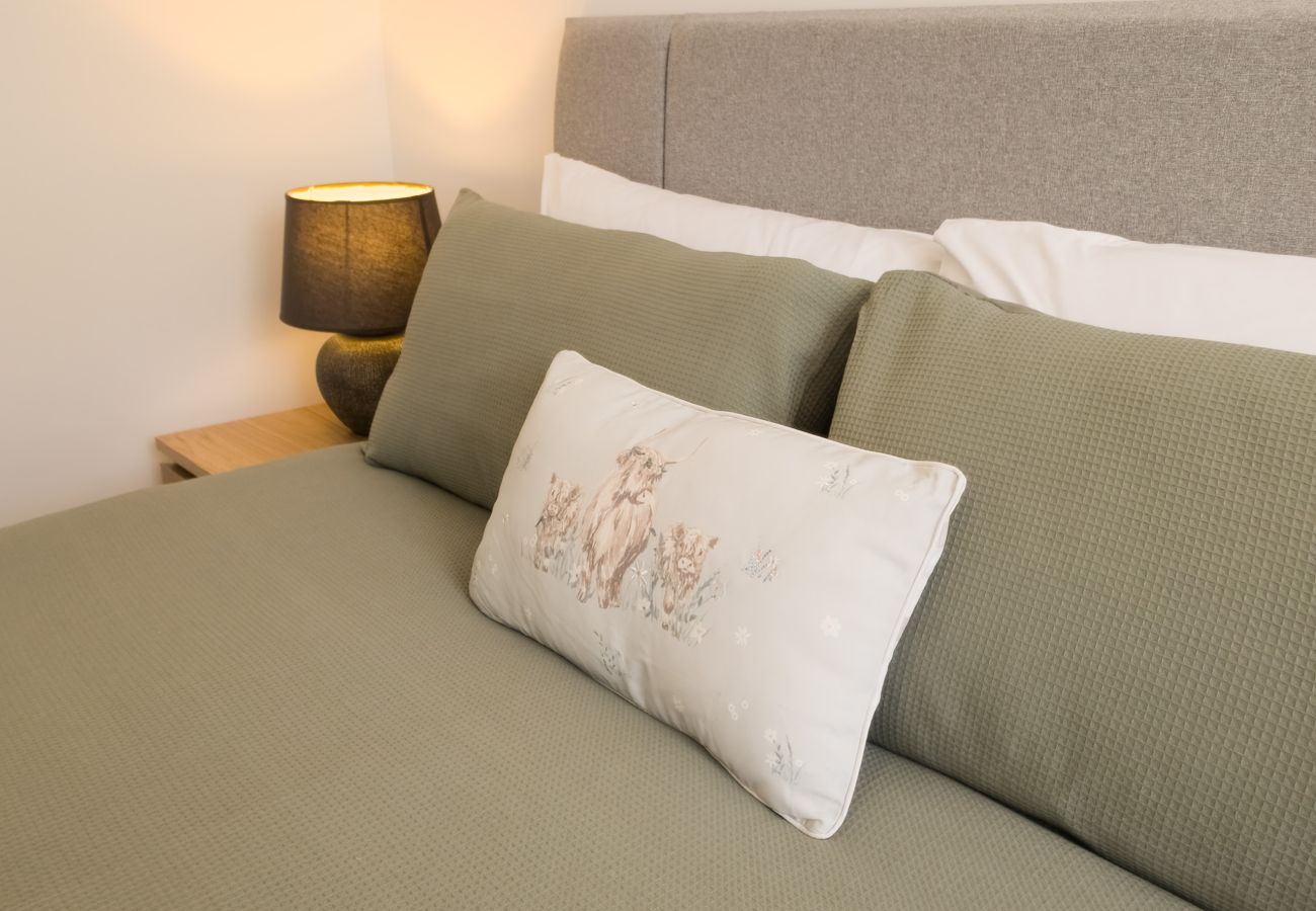 Close-up of a double bed with sage green pillows and an embroidered cushion, lit by a bedside lamp