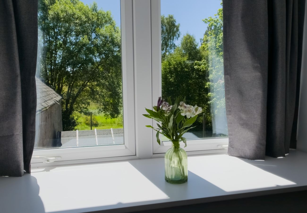 Bedroom window with grey curtains and a vase of white flowers looking out onto green trees in the sunshine