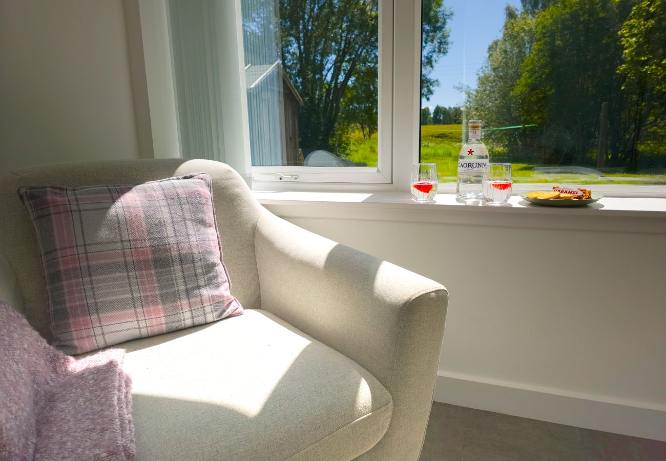 Cream armchair with a pink tartan cushion beside a sunny window looking out over green trees and lawn