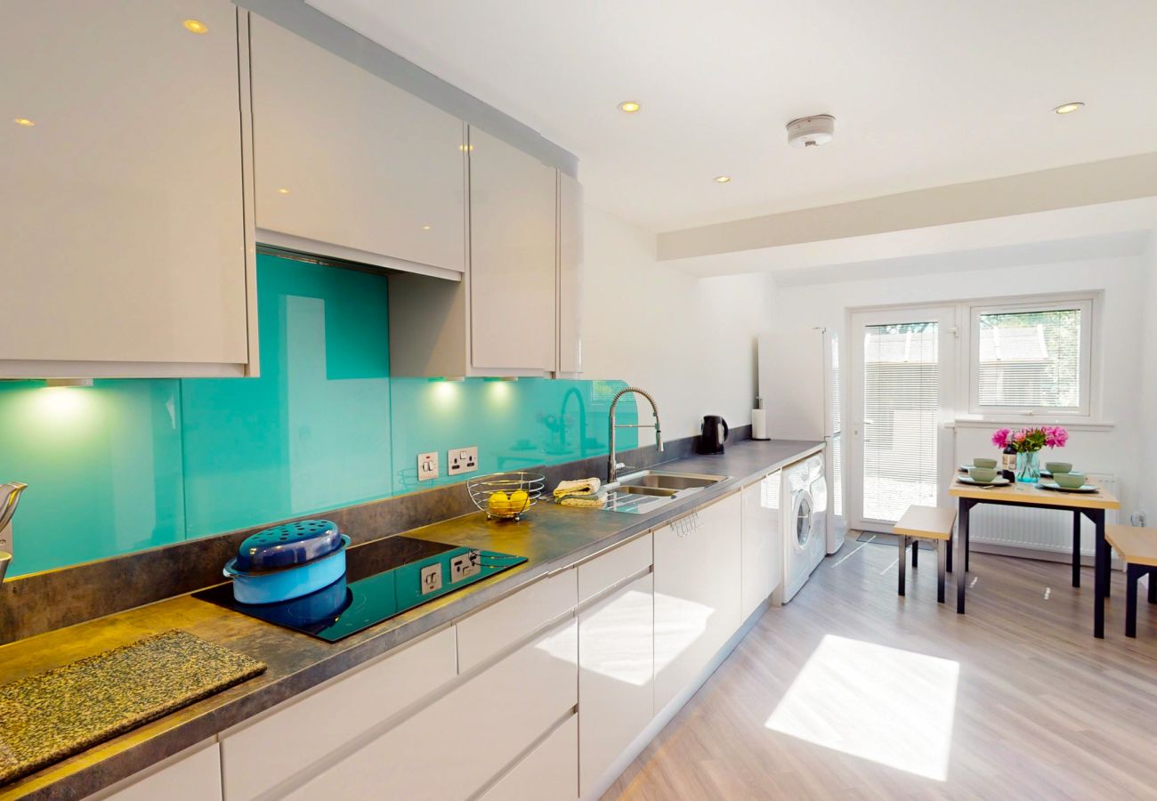 Long modern kitchen with turquoise splashback and washing machine, leading to a dining table by glazed doors