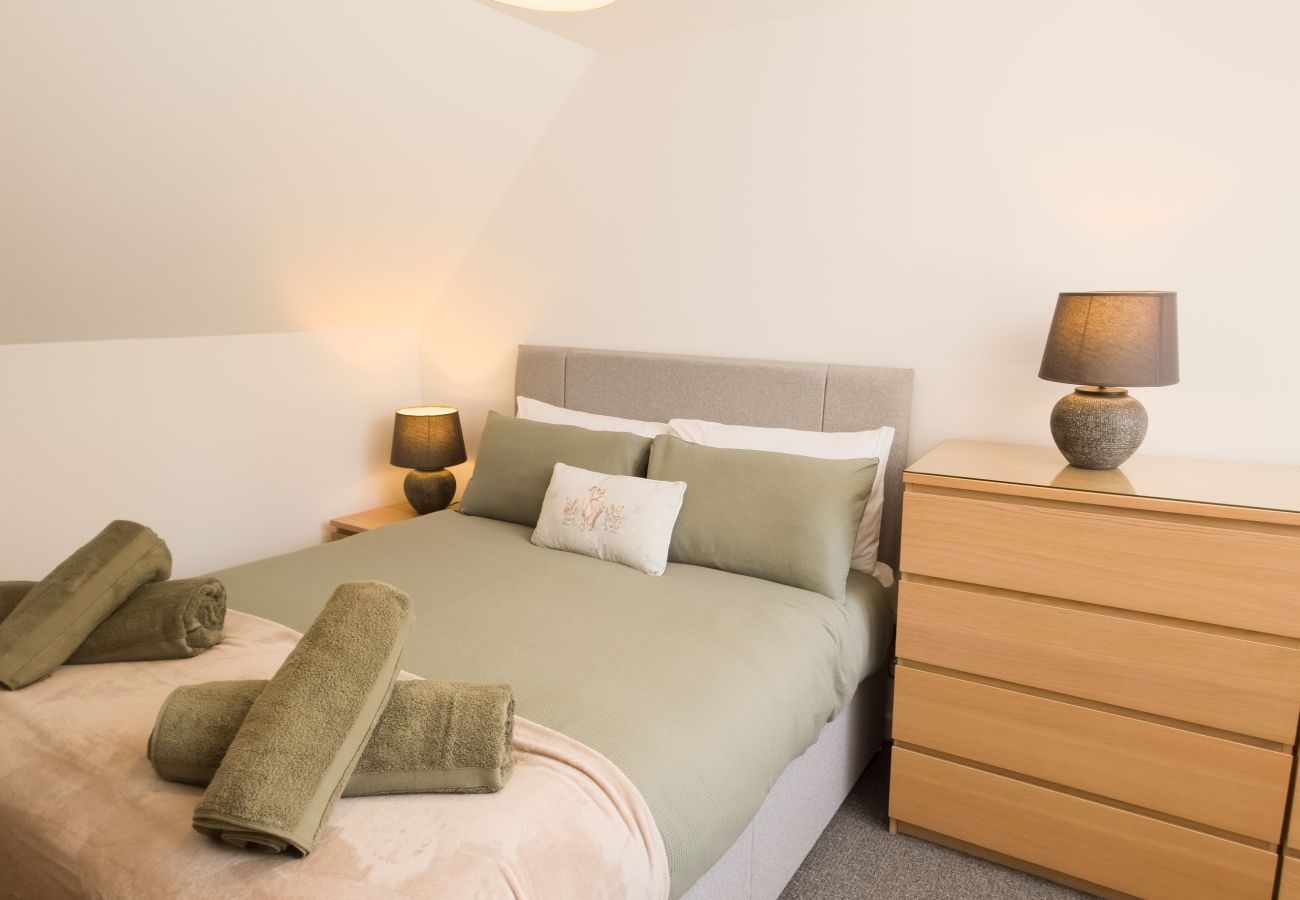 Double bed with sage green linen, rolled towels, bedside lamps and an oak chest of drawers in a bright bedroom